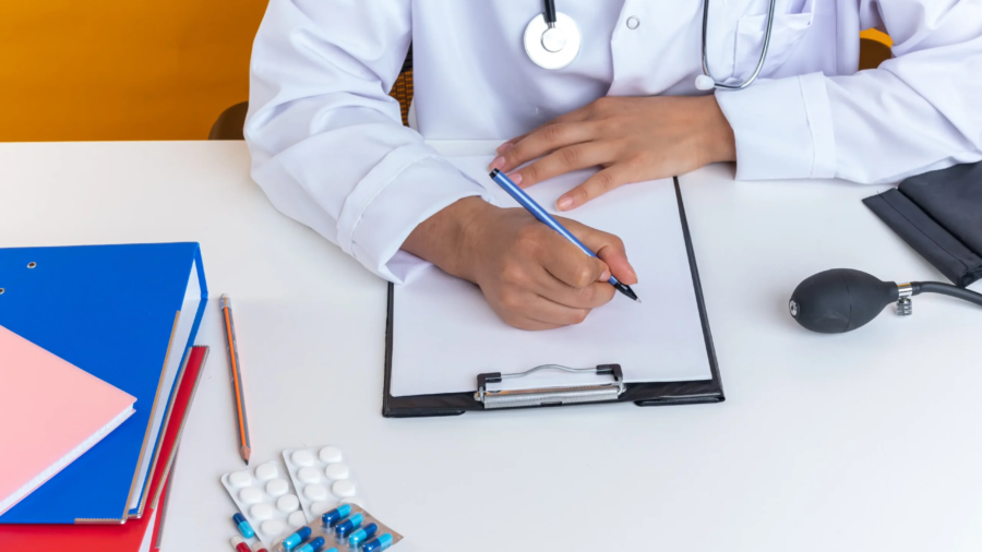 young-female-doctor-wearing-medical-robe-with-stethoscope-sits-table-with-medical-tools-writing-something-clipboard-isolated-yellow-background